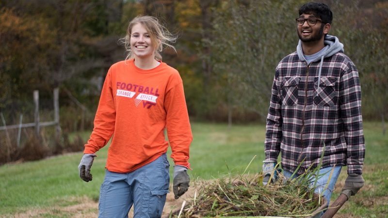 Student pushes wheelbarrel full of weeds as second student walks alongside.